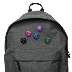 Op de afbeelding: Een grijze rugzak met vijf ronde knopen bevestigd. De knopen zijn in verschillende kleuren, waaronder roze, blauw, groen, paars en zwart. Elke knoop heeft witte tekst in een cirkelvormig patroon.