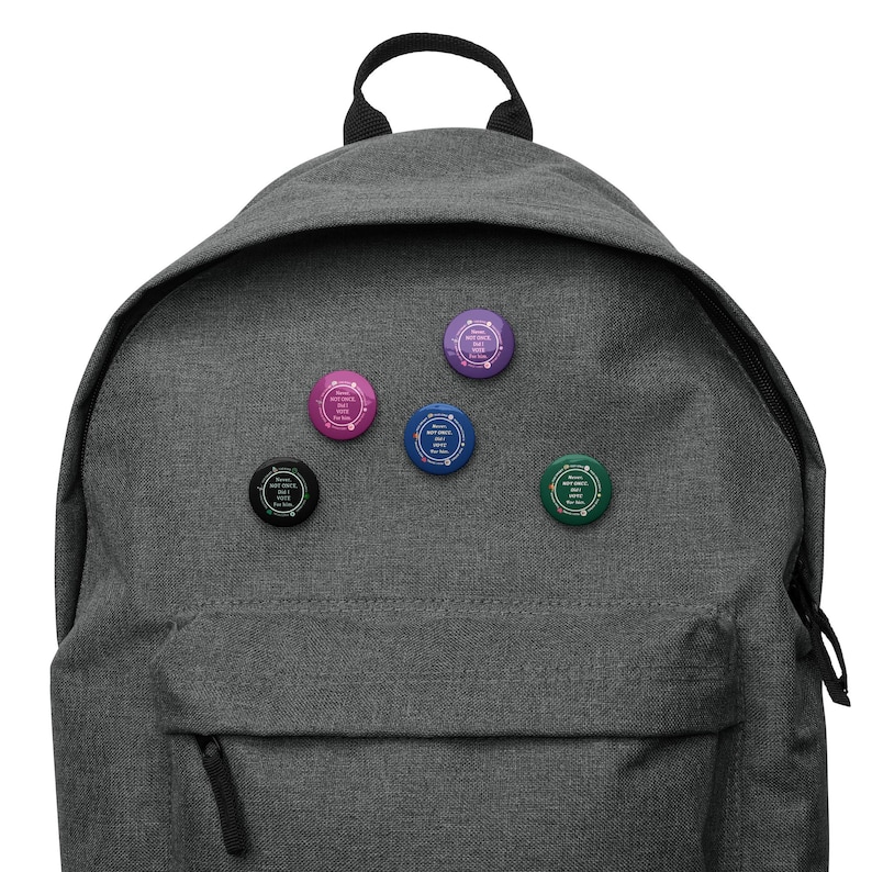 Op de afbeelding: Een grijze rugzak met vijf ronde knopen bevestigd. De knopen zijn in verschillende kleuren, waaronder roze, blauw, groen, paars en zwart. Elke knoop heeft witte tekst in een cirkelvormig patroon.