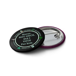 Op de afbeelding: Een zwarte button met de tekst "Never, NOT ONCE, Did I VOTE For him." en zinnen als "Too Smart" en "Too Human." Het heeft een zilveren speld en een paarse rand. Een politieke uitspraak.
