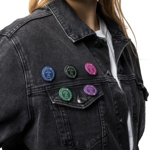Op de afbeelding: Een zwarte spijkerjas met zes ronde pins. Elke pin toont de tekst "Never Not Once But Vote For Him" in het wit, op blauwe, groene, roze en paarse achtergronden. De jas wordt gedragen over een wit shirt.