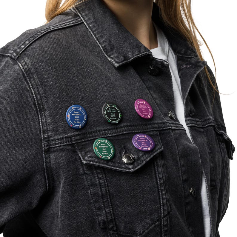 Op de afbeelding: Een zwarte spijkerjas met zes ronde pins. Elke pin toont de tekst "Never Not Once But Vote For Him" in het wit, op blauwe, groene, roze en paarse achtergronden. De jas wordt gedragen over een wit shirt.