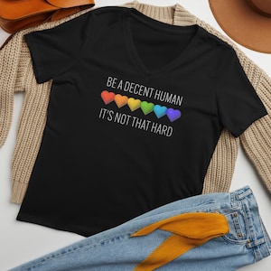 May include: Black V-neck t-shirt with the text "BE A DECENT HUMAN IT'S NOT THAT HARD" and a rainbow heart design. The shirt is displayed with blue jeans, a tan sweater, and a brown hat.