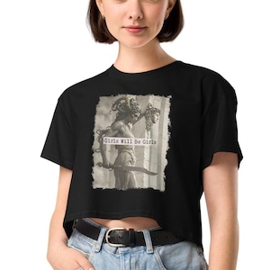 Woman wearing a black cropped T-shirt with a graphic of Medusa holding a sword and a decapitated head, featuring the text ‘Girls Will Be Girls’; styled with light blue high-waisted jeans and a black belt.