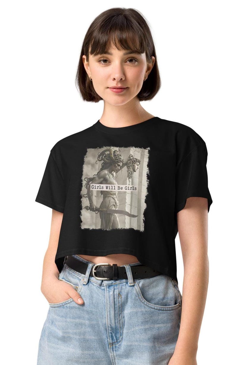 Woman wearing a black cropped T-shirt with a graphic of Medusa holding a sword and a decapitated head, featuring the text ‘Girls Will Be Girls’; styled with light blue high-waisted jeans and a black belt.