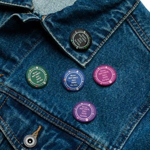 Op de afbeelding: Een verzameling ronde buttons in verschillende kleuren, waaronder groen, blauw, paars en zwart, zijn vastgemaakt aan een spijkerjasje. Elke button bevat de tekst "Never, NOT ONCE, Did I VOTE For him." in een cirkelvormig ontwerp.