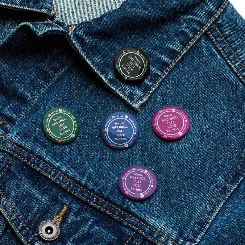 Op de afbeelding: Een verzameling ronde buttons in verschillende kleuren, waaronder groen, blauw, paars en zwart, zijn vastgemaakt aan een spijkerjasje. Elke button bevat de tekst "Never, NOT ONCE, Did I VOTE For him." in een cirkelvormig ontwerp.