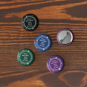 Op de afbeelding: Vijf ronde buttons met de tekst "Never, NOT ONCE, Did I VOTE For him." in het wit op verschillende gekleurde achtergronden: zwart, blauw, groen, paars en een roze achterkant. De buttons liggen op een houten oppervlak.