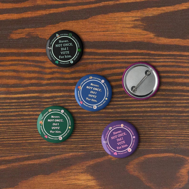 Op de afbeelding: Vijf ronde buttons met de tekst "Never, NOT ONCE, Did I VOTE For him." in het wit op verschillende gekleurde achtergronden: zwart, blauw, groen, paars en een roze achterkant. De buttons liggen op een houten oppervlak.