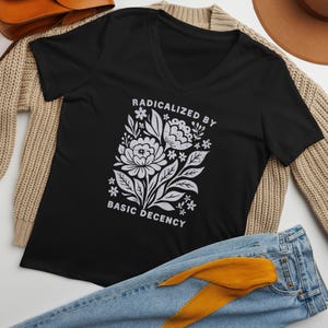 May include: Black V-neck t-shirt with a white floral design and the text "RADICALIZED BY BASIC DECENCY". The shirt is displayed on a neutral-colored sweater and blue jeans.
