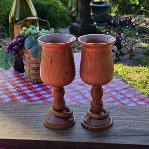 Puede incluir: Dos copas de vino de madera con anillos alrededor de los tallos, sentadas sobre una tabla de madera. Las copas son de color marrón claro y tienen un acabado liso y pulido.
