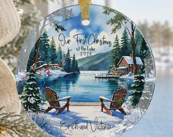 Puede incluir: Un adorno de cristal redondo con una escena de lago invernal que incluye dos sillas de madera, una cabaña y árboles de hoja perenne. El texto dice Our First Christmas at the Lake 2026 y Erich and Victoria. Cuelga de una cinta dorada.