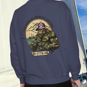 May include: Navy blue sweatshirt featuring a graphic of a Bigfoot-like figure fishing, wearing a hat with an American flag design. The graphic includes the text "250 YEARS OF AMERICAN ANGLING" and "1776". The design is set against a mountain and lake backdrop.