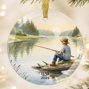 May include: A round glass ornament featuring a watercolor illustration of a person in a hat and blue shirt fishing from a log by a lake at sunset. The text LEVI 2025 is printed in black at the bottom. A gold ribbon loop is attached to the top.