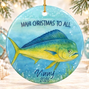 May include: A round ceramic Christmas ornament featuring a vibrant illustration of a mahi-mahi fish swimming in blue water. The text "MAHI CHRISTMAS TO ALL" is printed at the top, with the name "VINNY" and the year "2026" printed at the bottom.