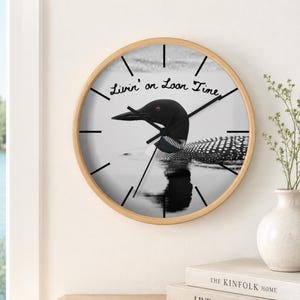 May include: A round wall clock with a light wood frame featuring a black and white photograph of a loon swimming on water. The clock face displays the text Livin' on Loon Time in black script, with black hour and minute hands and simple markers.