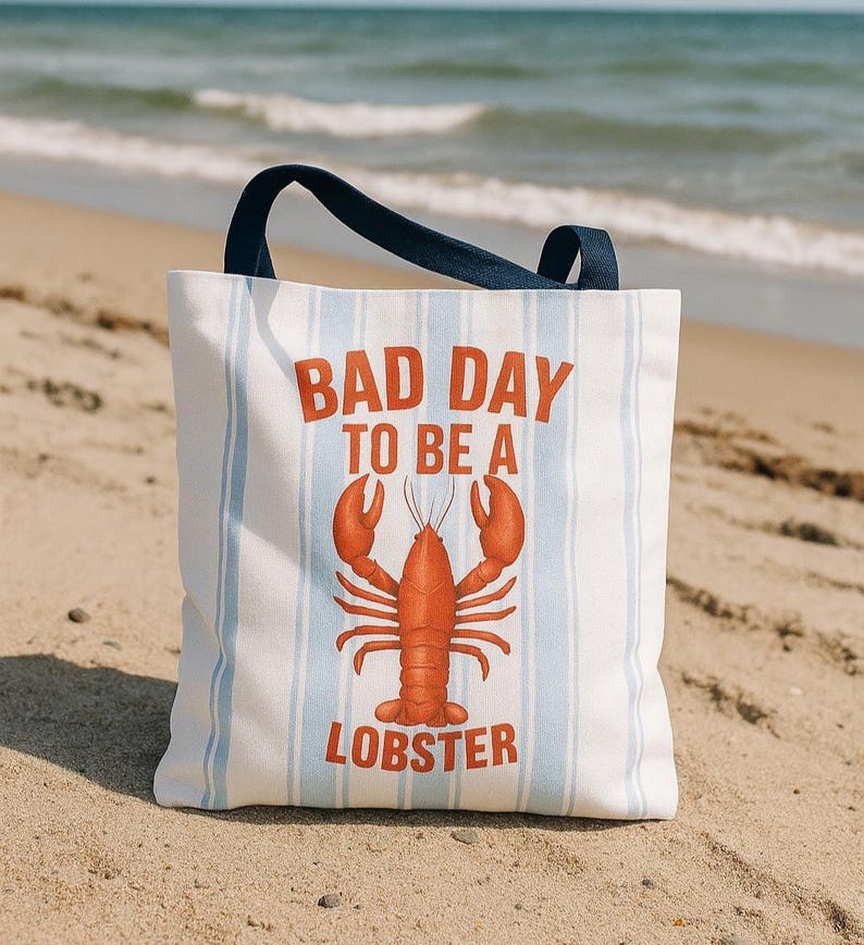 Peut inclure: Un sac fourre-tout blanc avec des rayures verticales bleues et une poign&eacute;e bleu marine. Le sac affiche le texte "BAD DAY TO BE A LOBSTER" en orange, avec une illustration de homard orange. Le sac est sur une plage de sable.
