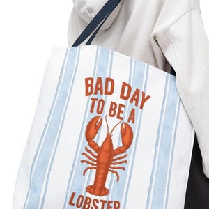 Peut inclure: Sac fourre-tout blanc avec des rayures verticales bleues et une illustration de homard rouge. Le texte "BAD DAY TO BE A LOBSTER" est imprim&eacute; en rouge. Le sac a une sangle bleu marine.