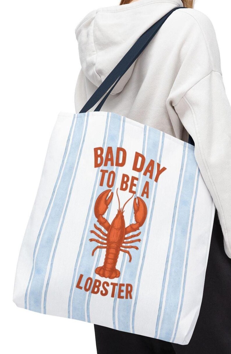 Peut inclure: Sac fourre-tout blanc avec des rayures verticales bleues et une illustration de homard rouge. Le texte "BAD DAY TO BE A LOBSTER" est imprim&eacute; en rouge. Le sac a une sangle bleu marine.