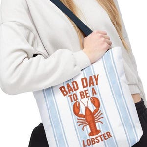 Peut inclure: Sac fourre-tout blanc &agrave; rayures verticales bleues et une sangle bleu marine. Le sac porte l'inscription "BAD DAY TO BE A LOBSTER" en rouge, ainsi qu'une illustration de homard rouge. Le sac est port&eacute; &agrave; l'&eacute;paule.