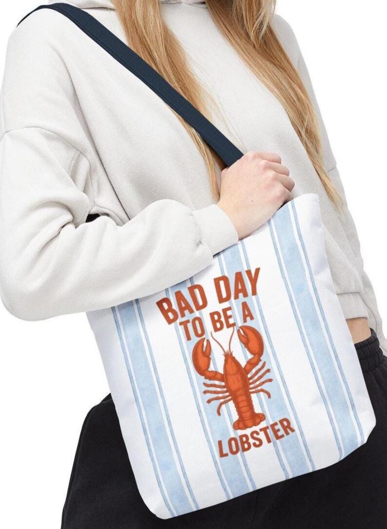 Peut inclure: Sac fourre-tout blanc &agrave; rayures verticales bleues et une sangle bleu marine. Le sac porte l'inscription "BAD DAY TO BE A LOBSTER" en rouge, ainsi qu'une illustration de homard rouge. Le sac est port&eacute; &agrave; l'&eacute;paule.