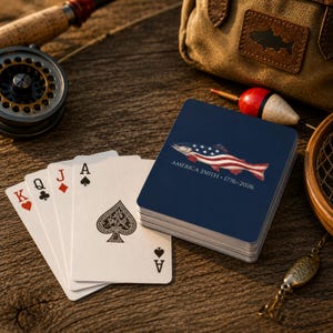 May include: A deck of playing cards with a dark blue back featuring a fish graphic patterned with the American flag and the text AMERICA 250TH 1776-2026. Four playing cards are fanned out beside the deck, showing the King of Hearts, Queen of Clubs, Jack of Diamonds, and Ace of Spades.