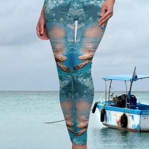 Op de afbeelding: Een blauwe legging met een zeeschildpad en oceaan golfpatroon. De legging wordt gedragen door een persoon die voor een boot in de oceaan staat.