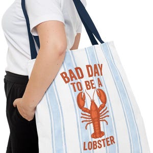 Peut inclure: Sac fourre-tout blanc &agrave; rayures verticales bleues et anses bleu marine. Le sac affiche le texte "BAD DAY TO BE A LOBSTER" en rouge, avec un dessin de homard rouge. Le sac est port&eacute; &agrave; l'&eacute;paule.
