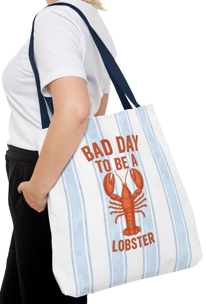 Peut inclure: Sac fourre-tout blanc &agrave; rayures verticales bleues et anses bleu marine. Le sac affiche le texte "BAD DAY TO BE A LOBSTER" en rouge, avec un dessin de homard rouge. Le sac est port&eacute; &agrave; l'&eacute;paule.