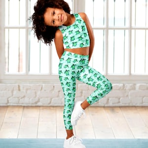 May include: A young girl poses in a matching turquoise athletic set. The set includes a crop top and leggings, both patterned with green sea turtles. She wears white sneakers and stands on a blue mat in front of a window.