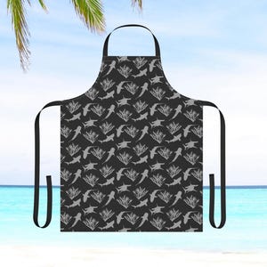 May include: Black apron featuring a white shark and coral reef design. The apron has adjustable neck straps and two long ties.