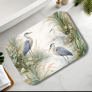 May include: Rectangular bath mat featuring a watercolor design of three herons in a marsh setting. The mat has a white background with green and beige foliage and white flowers. Two herons are blue and gray, and one is white.