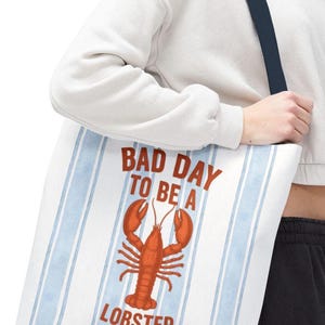 Peut inclure: Sac fourre-tout blanc avec des rayures verticales bleues et une illustration de homard rouge. Le texte sur le sac se lit "BAD DAY TO BE A LOBSTER". Le sac a une sangle bleu marine.