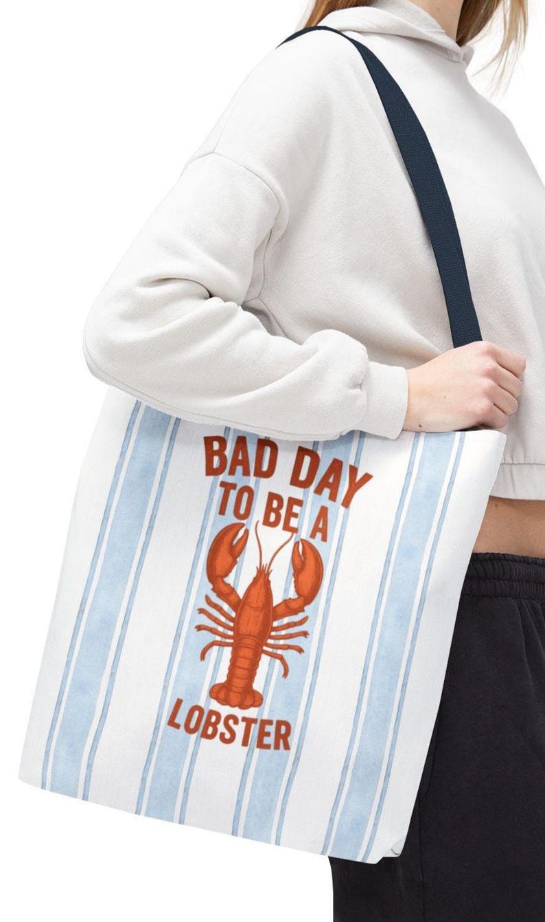 Peut inclure: Sac fourre-tout blanc avec des rayures verticales bleues et une illustration de homard rouge. Le texte sur le sac se lit "BAD DAY TO BE A LOBSTER". Le sac a une sangle bleu marine.