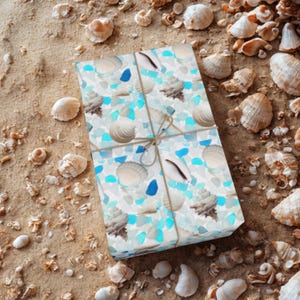 May include: A wrapped gift box with a seashell and sea glass pattern, tied with twine, rests on a sandy surface scattered with seashells. The gift wrap features white, blue, and tan colors.