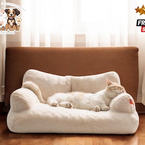 Puede incluir: Una cama para gatos de piel sintética blanca con forma de sofá con un gato durmiendo en ella. La cama tiene una pequeña almohada en un extremo.
