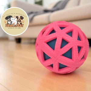 Puede incluir: Una pelota rosa para perros con un diseño geométrico, con secciones interiores azules. La pelota está sobre un suelo de madera, con un fondo borroso de un sofá y un logotipo que dice "Paws & Play".