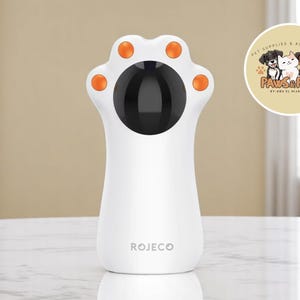 Peut inclure: Un jouet pour chat blanc en forme de patte, avec des détails orange. Le centre est noir et le mot "ROJECO" est imprimé sur la base. Un logo "Paws & Play" est visible en arrière-plan. Ce produit est conçu pour les animaux de compagnie.
