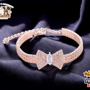 May include: A rose gold dog collar featuring a sparkling bow with a clear rhinestone at its centre. The collar is made of metal and has a chain link design.