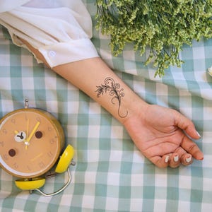 May include: A yellow alarm clock with black numbers and hands on a green and white checkered surface. The clock is set to 9:00. A person's arm with a small floral tattoo is visible in the background.