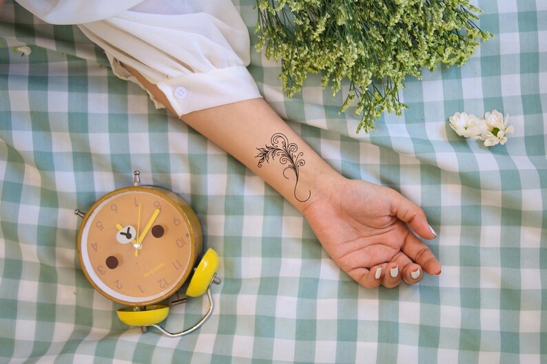 May include: A yellow alarm clock with black numbers and hands on a green and white checkered surface. The clock is set to 9:00. A person's arm with a small floral tattoo is visible in the background.