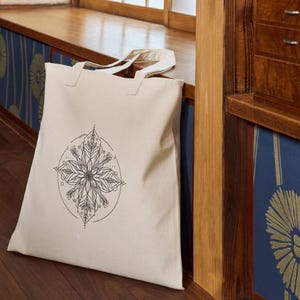 May include: A white canvas tote bag with a black and white mandala design. The mandala is a circular design with intricate details and floral patterns.