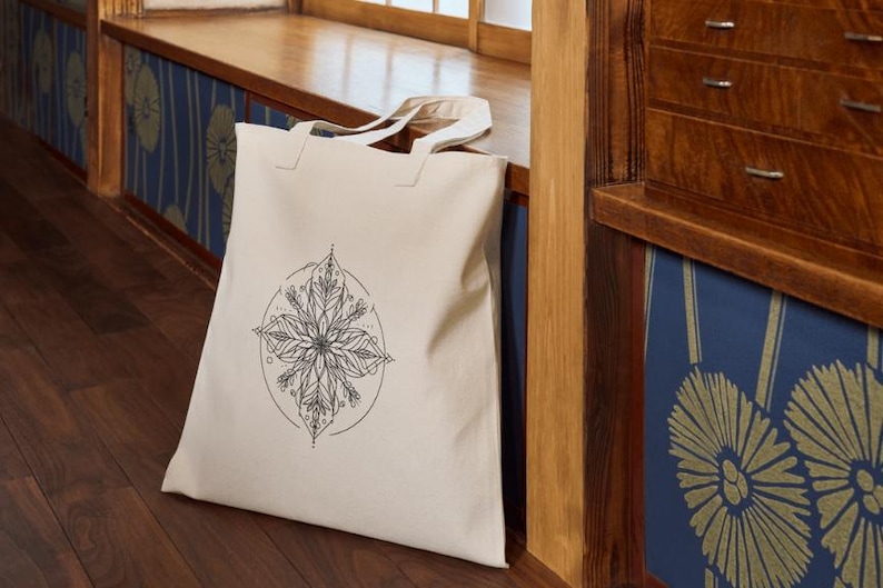 May include: A white canvas tote bag with a black and white mandala design. The mandala is a circular design with intricate details and floral patterns.