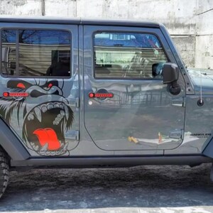 May include: A gray Jeep Wrangler with a black top and a large gorilla graphic on the side. The gorilla graphic is black, gray, and red. The Jeep has black side steps and red accents on the door handles.