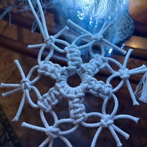 May include: A white macrame snowflake ornament with a six-pointed star design. The snowflake is made with thick white rope and has a knotted fringe.