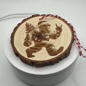 Op de afbeelding: Een rond houten ornament met schorsranden met een lasergravure van een Bigfoot-figuur met een kerstmuts en zonnebril, die een kerstboom draagt en een handgebaar maakt. Een rood-wit gedraaid touwtje is bevestigd.