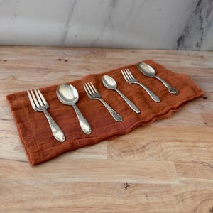 May include: Six pieces of vintage silverware, including forks and spoons, are arranged on a rust-colored linen napkin. The silverware has ornate handle detailing. The napkin rests on a light brown wooden surface, with a marble background.