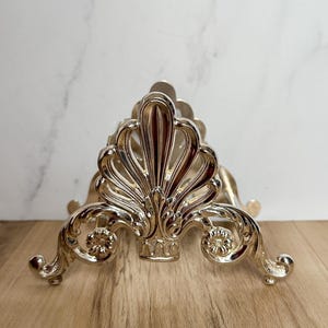 May include: Silver napkin holder with an ornate design. The holder has a fan-shaped top and intricate scrollwork on the sides.