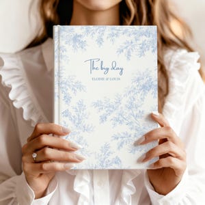 May include: A wedding journal with a blue floral pattern and the words "The big day" in script. The names "Elodie & Louis" are printed below. The journal is held by a person, with a white shirt in the background.