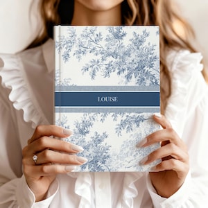 May include: A hardcover journal with a blue and white floral design. The journal features a navy blue band with the name "LOUISE" in white lettering. The cover has a toile pattern of branches and leaves.
