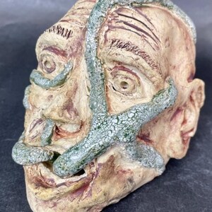 May include: A ceramic sculpture of a face with green vines growing over it. The face is textured and has a distressed look. The vines are detailed and have a speckled glaze.